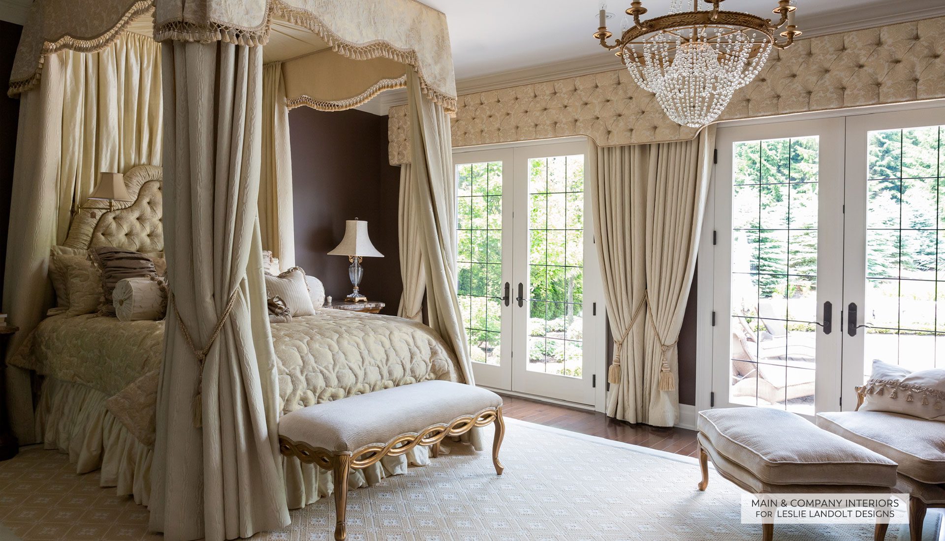 Elegant bedroom with a canopy bed, tufted headboard, ornate bench, chandelier, and French doors leading to a patio; decorated in cream and gold tones.