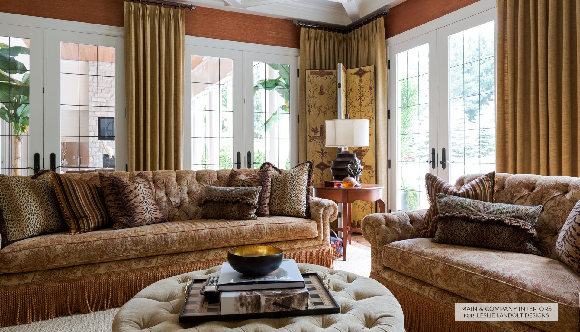 Two patterned sofas with decorative pillows face each other in a bright living room with large windows, gold curtains, and a tufted ottoman topped with a tray and decorative items.