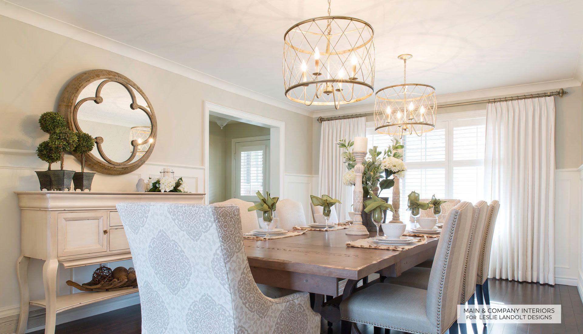 Elegant dining room with a wooden table, upholstered chairs, floral centerpieces, two chandeliers, a sideboard, and a round mirror on the wall. Sunlight streams through window drapes.