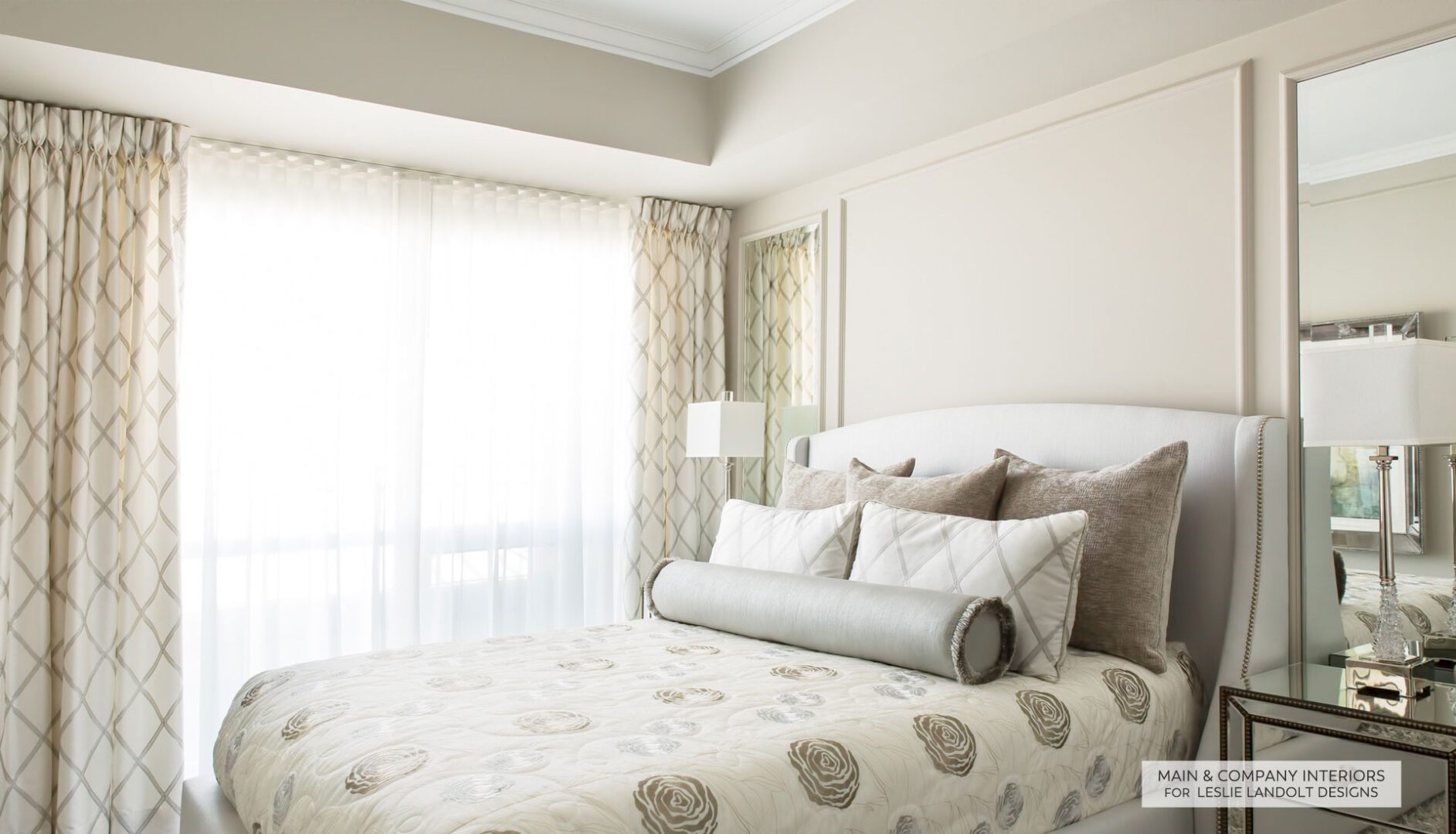 A bright bedroom with a large window, light-colored patterned curtains, an upholstered bed with neutral bedding, several decorative pillows, and a mirrored nightstand.