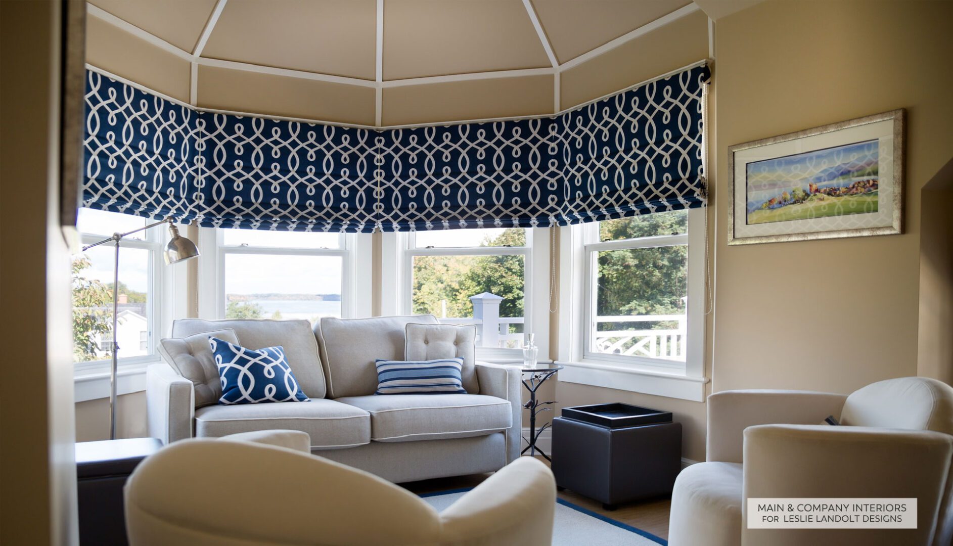 A cozy sunroom with large windows, a beige sofa with blue and white pillows, two armchairs, and patterned blue roman shades. A framed painting hangs on the tan wall.