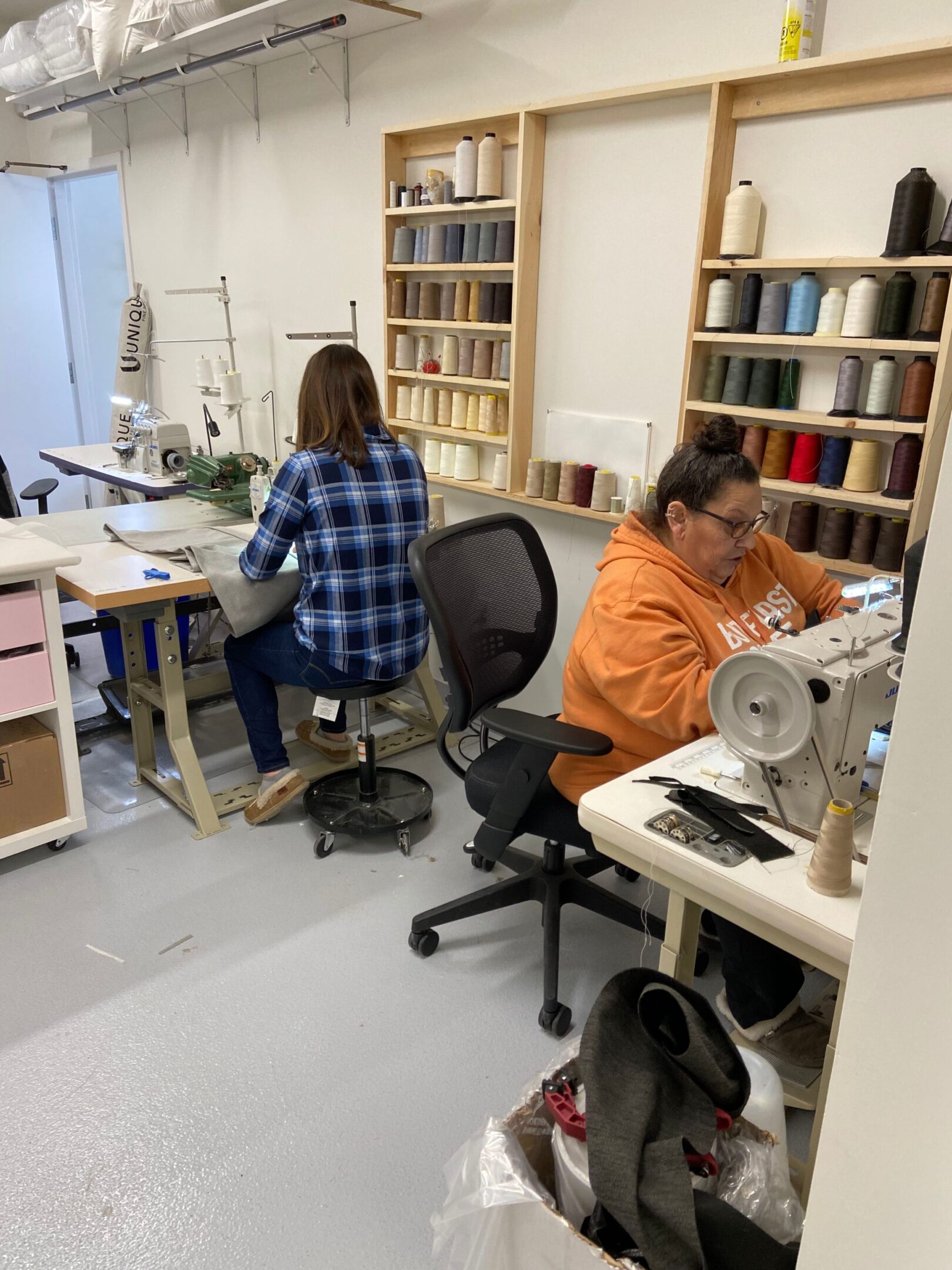 Two women working in a sewing workshop filled with fabric and shelves.