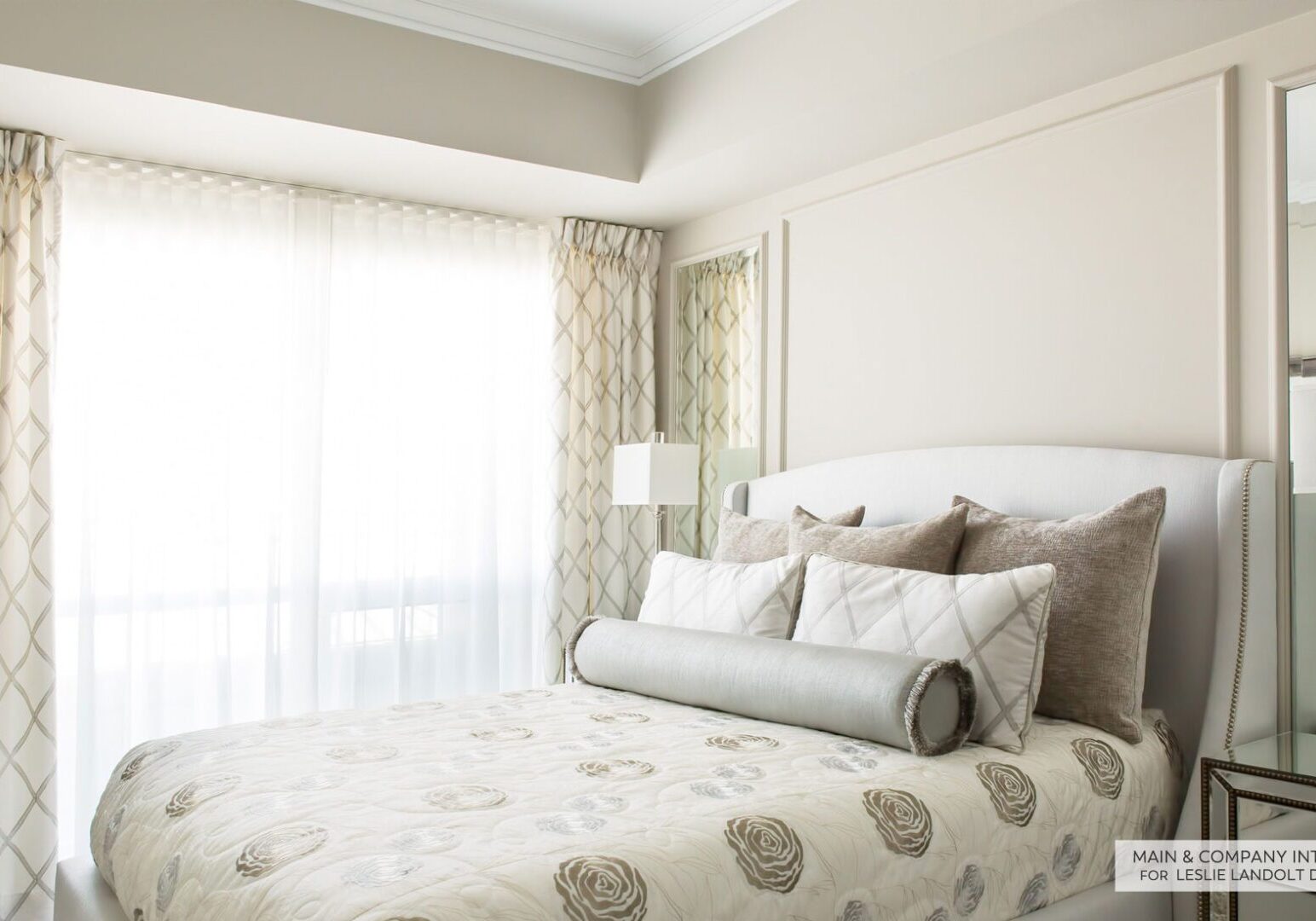 A bright bedroom with a large window, light-colored patterned curtains, an upholstered bed with neutral bedding, several decorative pillows, and a mirrored nightstand.