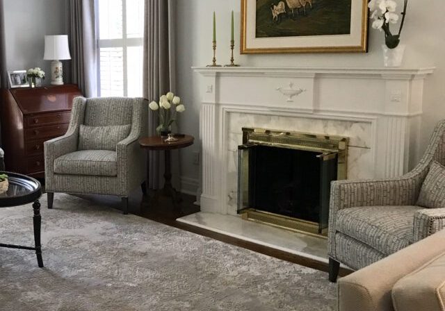 A living room with two armchairs, a fireplace with a brass screen, a round side table with white tulips, and a painting above the mantel.