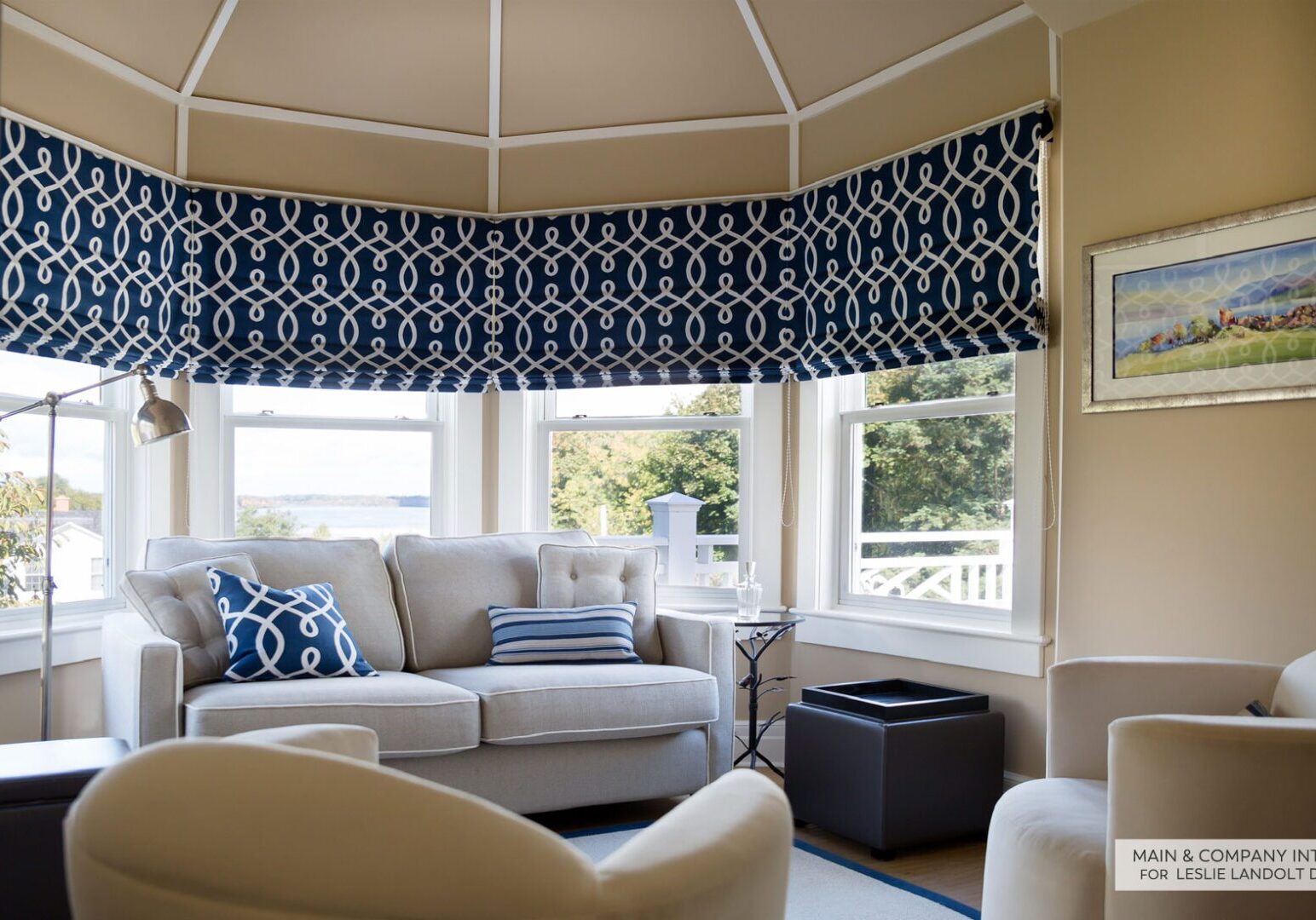A cozy sunroom with large windows, a beige sofa with blue and white pillows, two armchairs, and patterned blue roman shades. A framed painting hangs on the tan wall.